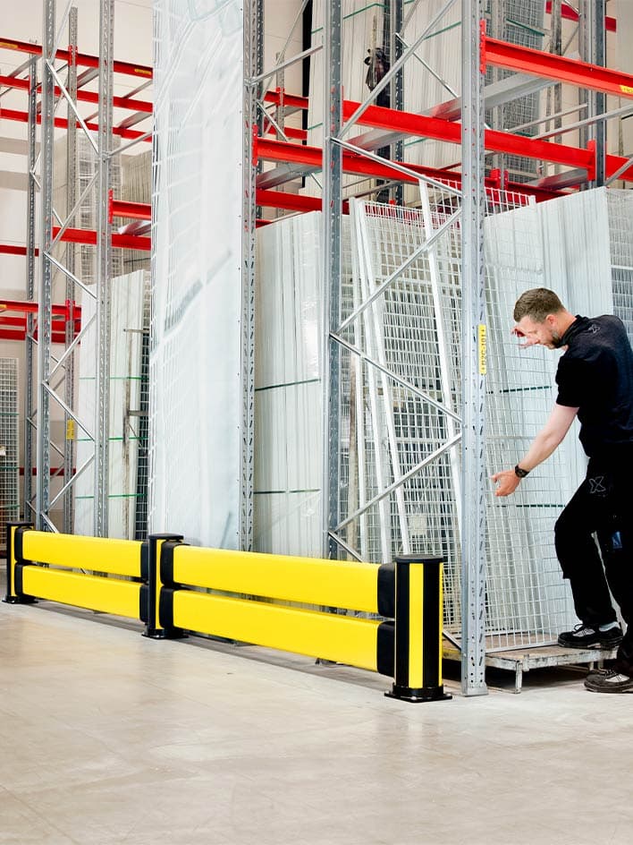 Warehouse worker handling glass panels behind yellow impact protection barriers. These industrial safety barriers prevent damage to fragile materials and protect employees from potential accidents, ensuring compliance with workplace safety regulations in manufacturing and storage environments.