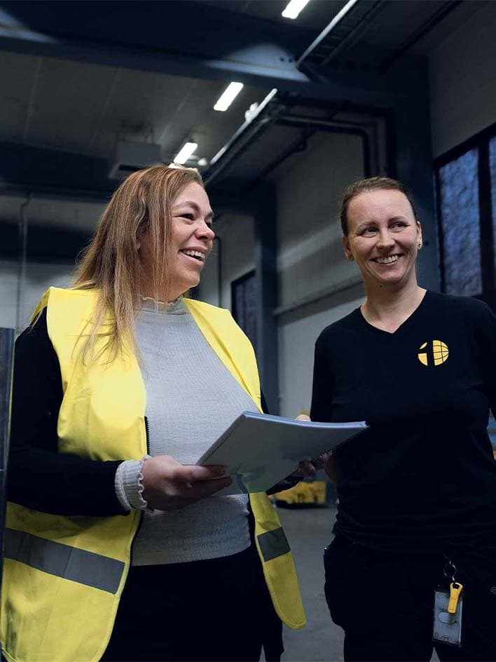 Two professionals in an industrial setting discussing new machinery regulations. One wears a high-visibility vest while holding compliance documents, emphasizing workplace safety, regulatory updates, and the transition to Machinery Regulation (2023/1230), replacing the Machinery Directive by January 20, 2027.
