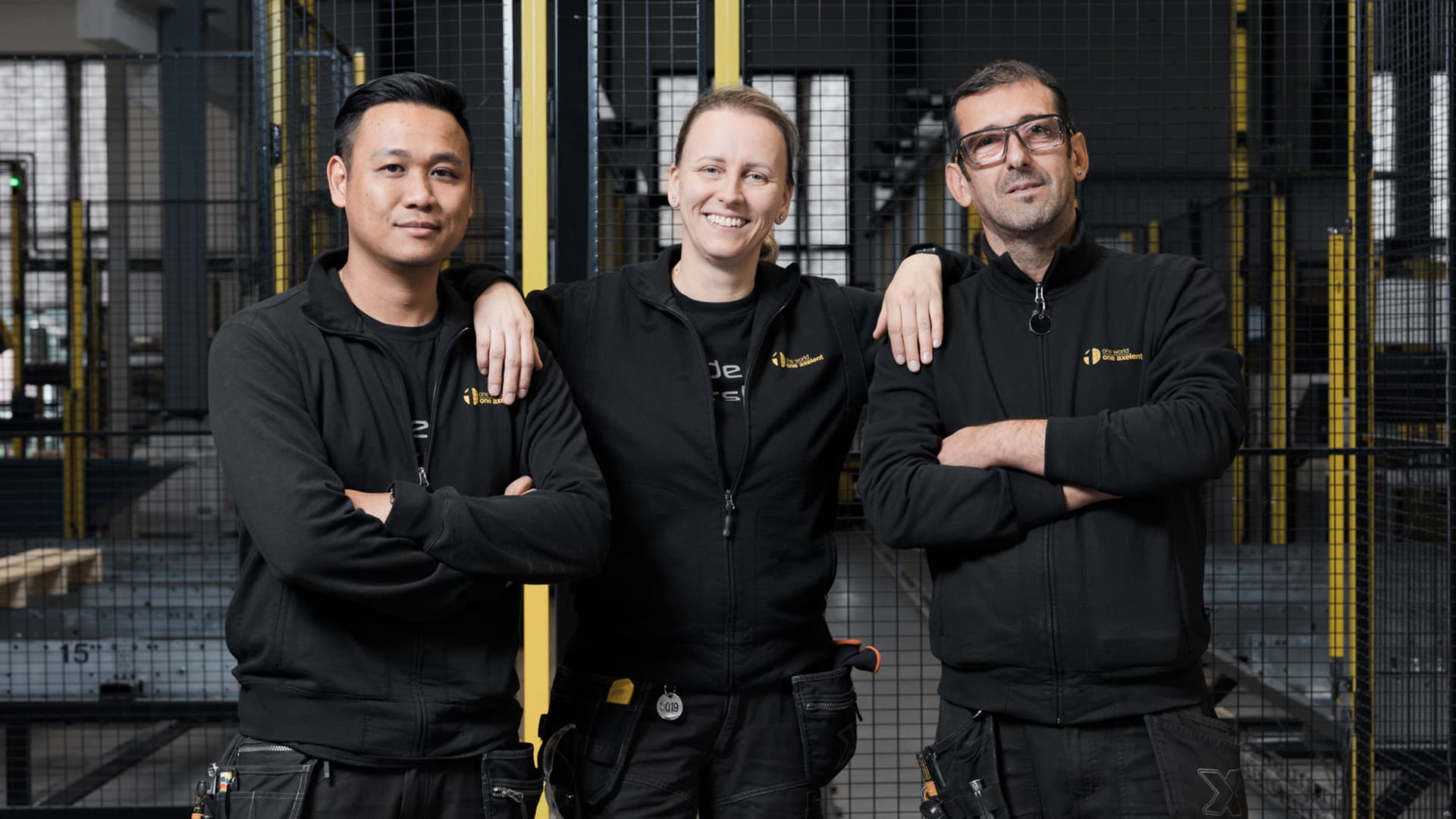Two men with crossed arms and a woman in the middle resting arms on each in front of machine guarding, all looking into the camera 