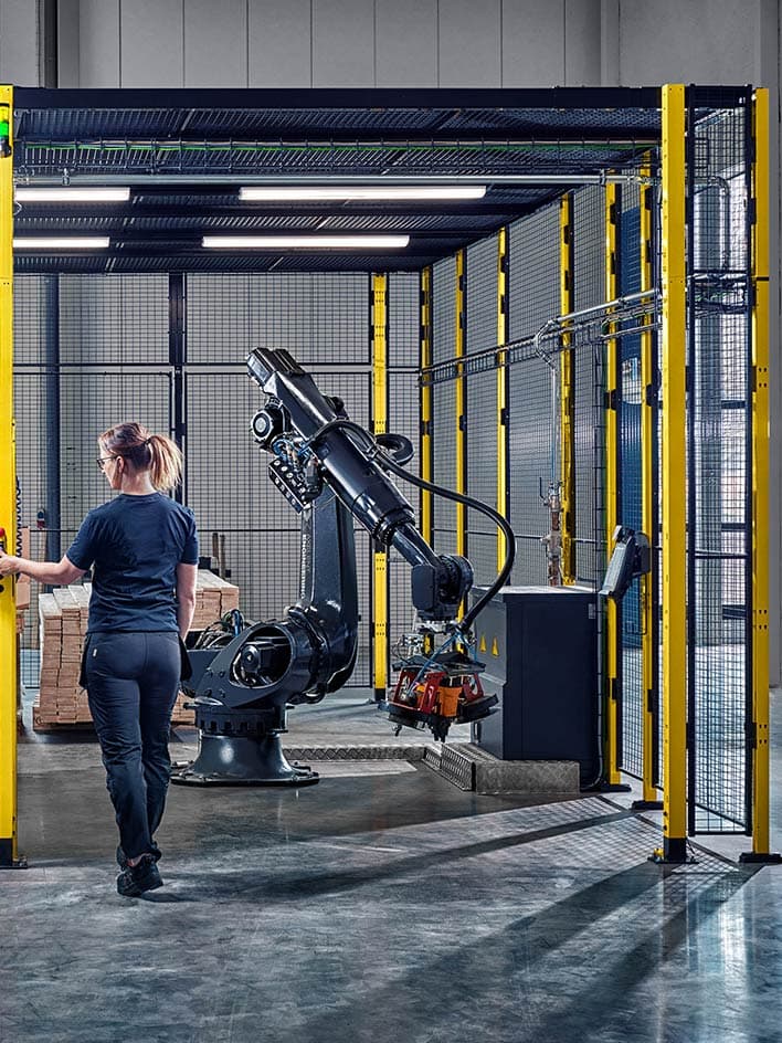 A worker enters a secure industrial robot cell, highlighting robotic safety measures, protective fencing, and access control in automated manufacturing environments. The image illustrates compliance with machine safety regulations and best practices for human-robot collaboration.