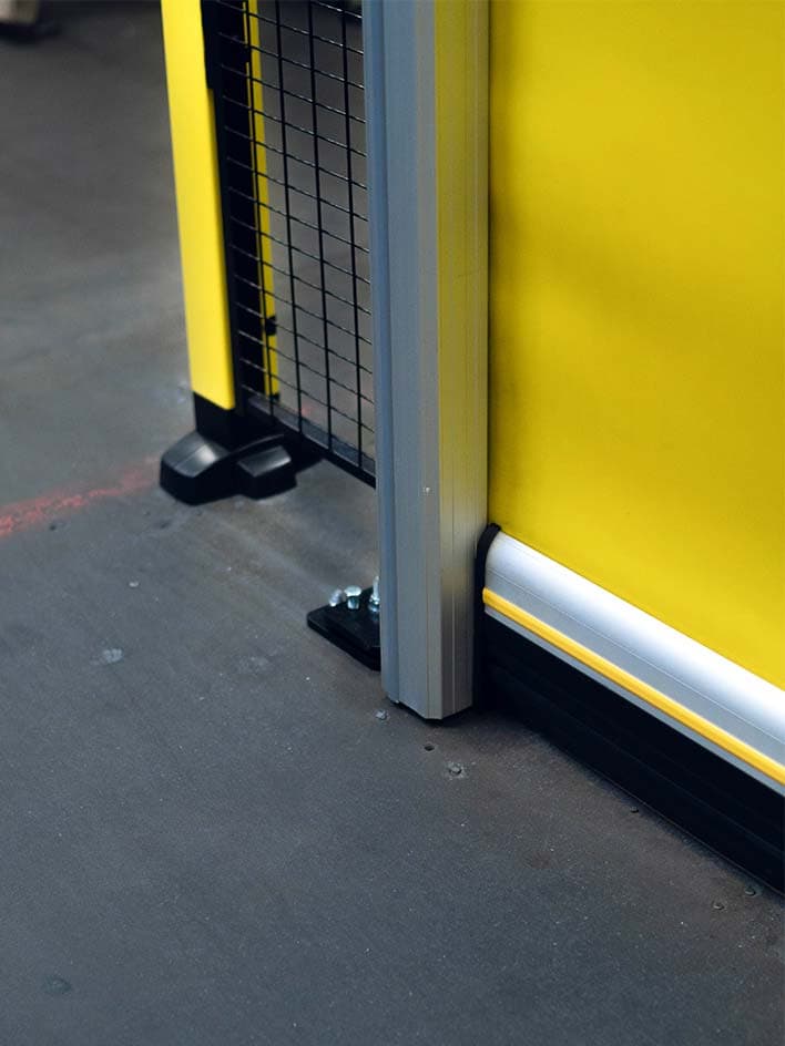Close-up of an industrial safety setup featuring guard fencing, a trip bar, and a protective bumper along the base of a yellow safety door. These components work together to detect contact and prevent hazards, ensuring a safer monitored access system