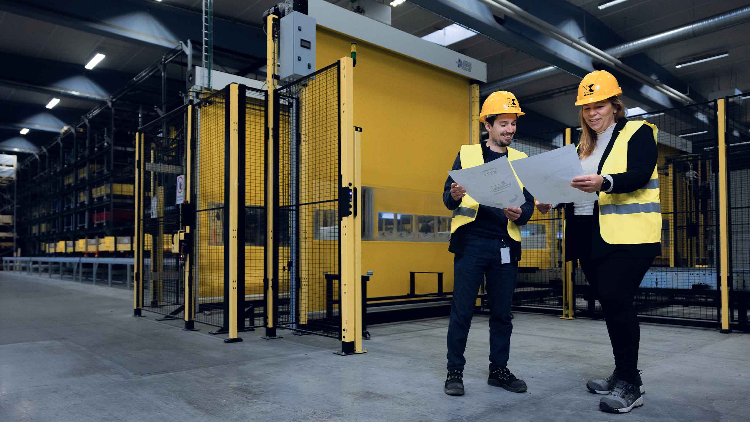 Two workers in safety vests and helmets reviewing technical documents in front of an automated roll-up door with guard fencing. The image highlights the role of monitoring systems in ensuring controlled access to industrial machinery as an alternative to movable guards.