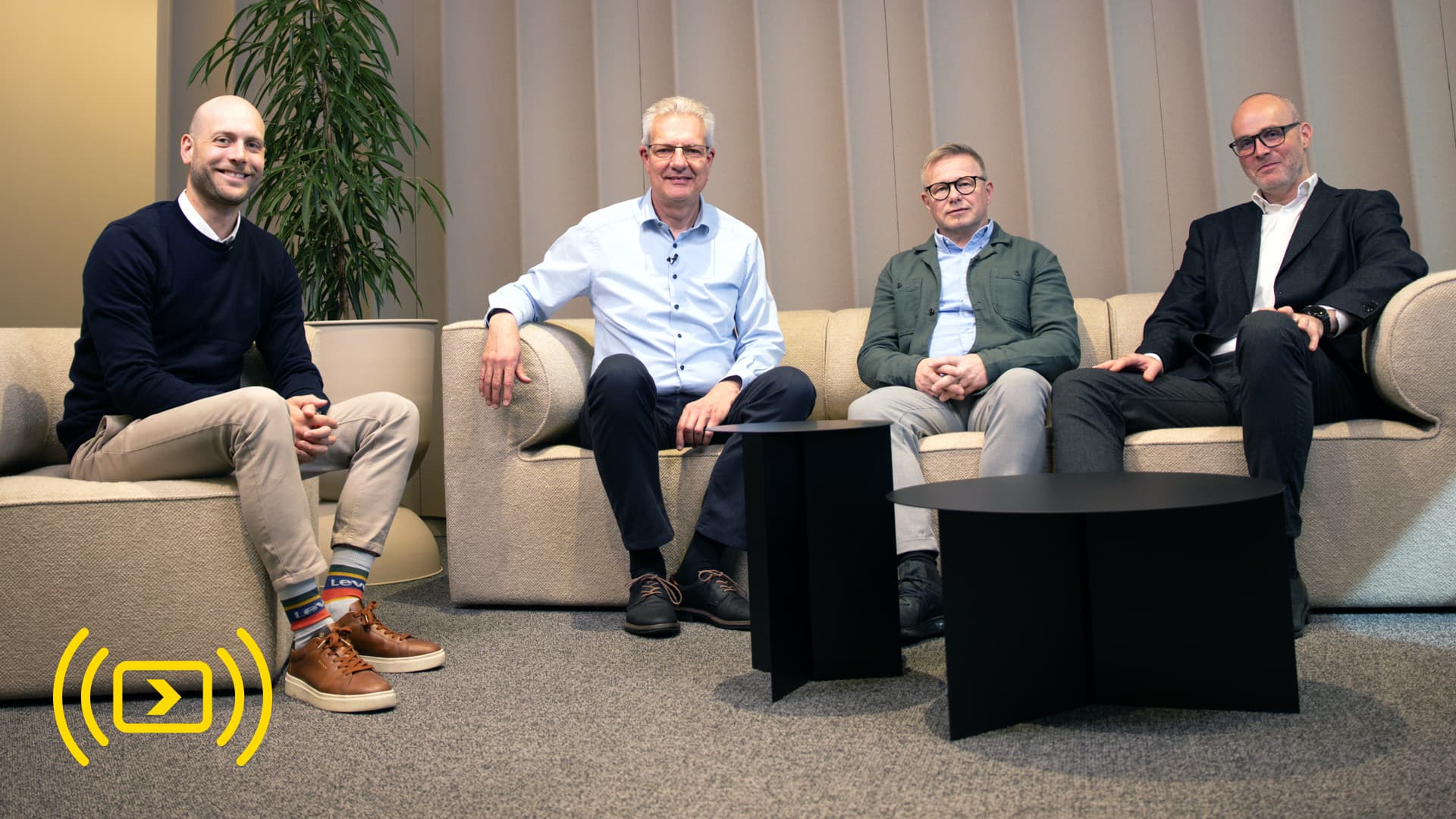 Four men sitting on a modern sofa in a studio setting, participating in a panel discussion or webinar.