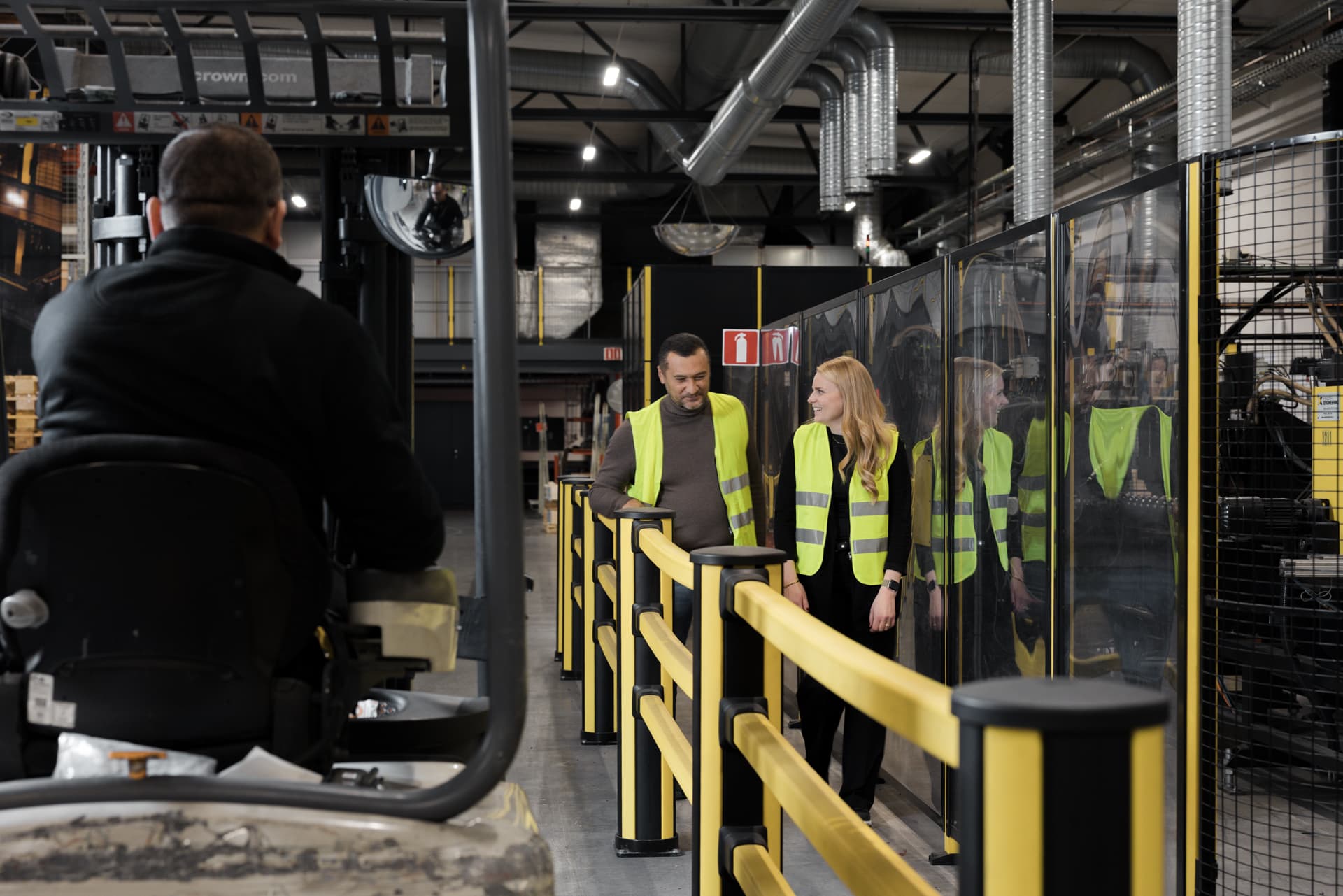 Two pedestrians are seen walking behind impact barriers while a forklift passes them in an industrial environment