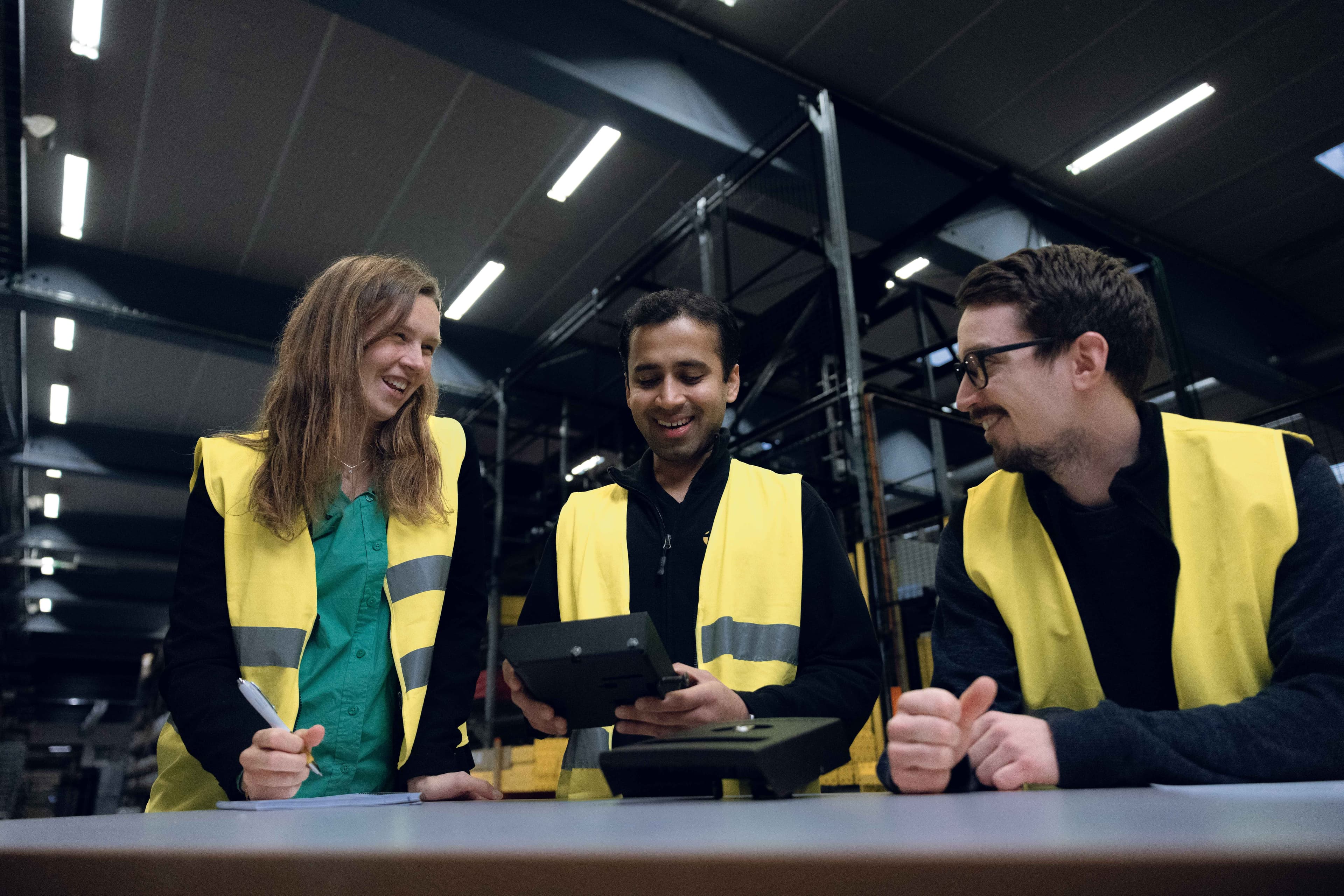 three people are shown in high visibility vests talking and laughing together with a warehouse in the background