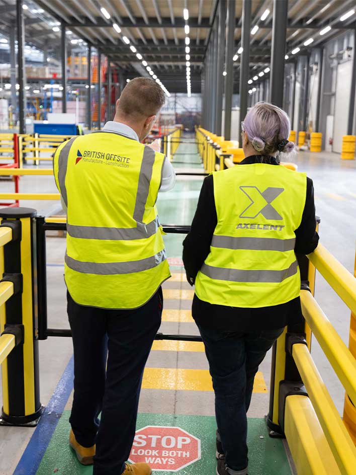 Two industrial safety professionals wearing high-visibility vests inspect a secure factory walkway, discussing global machinery safety standards and compliance regulations. The image highlights workplace safety measures, protective barriers, and risk management in industrial environments.