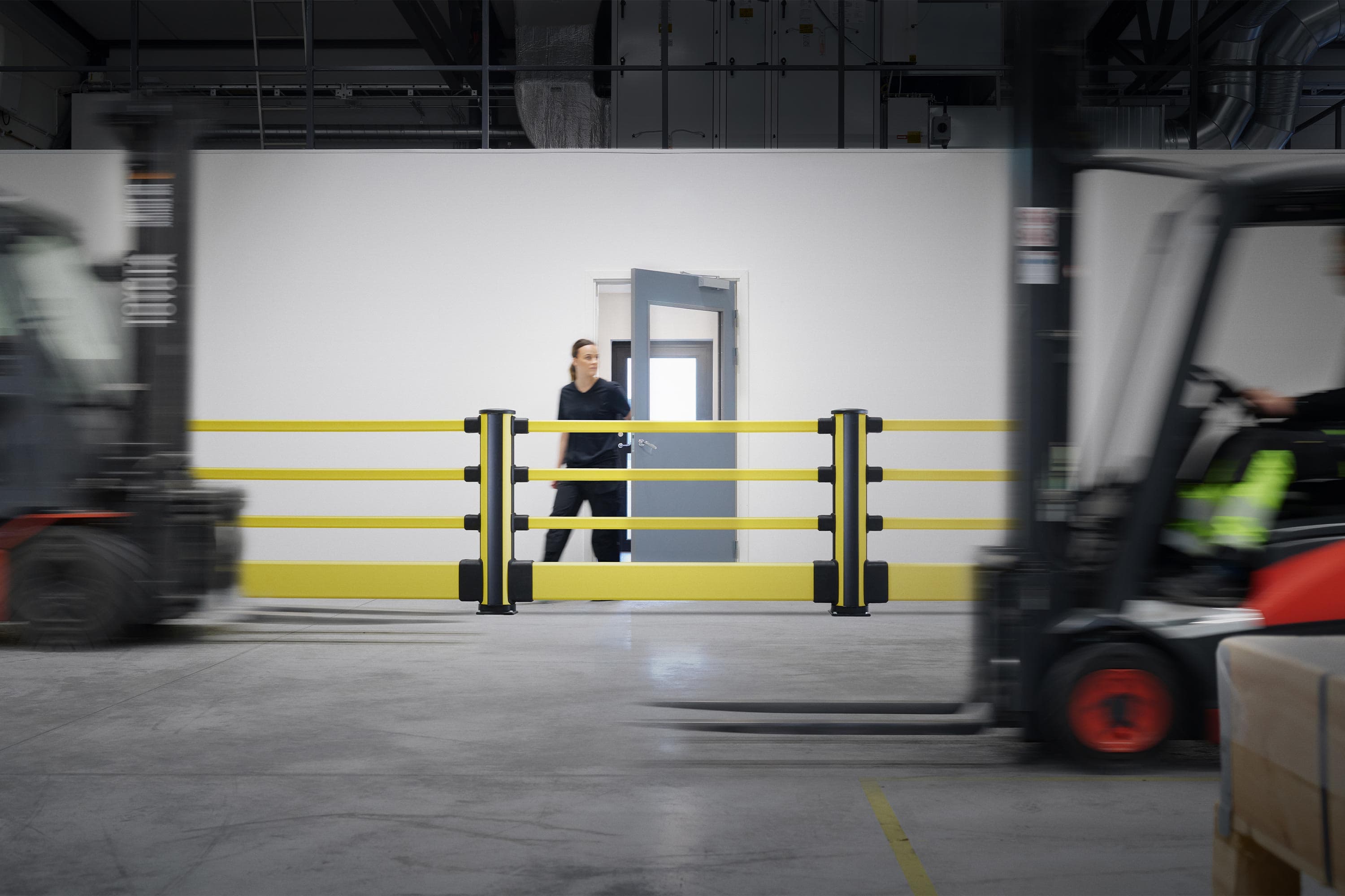 Pedestrian exiting a door protected by yellow safety barriers inside a warehouse, with a forklift passing