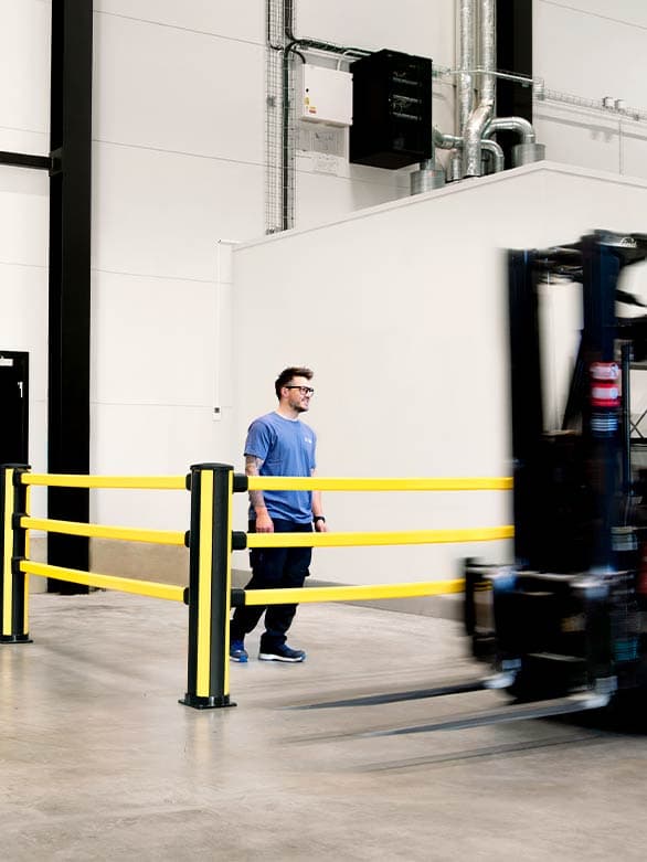 A pedestrian in an industrial warehouse walks safely behind a yellow crash barrier as a forklift passes by. The safety barrier acts as impact protection, preventing workplace accidents and ensuring compliance with industrial safety regulations for pedestrian and vehicle separation.