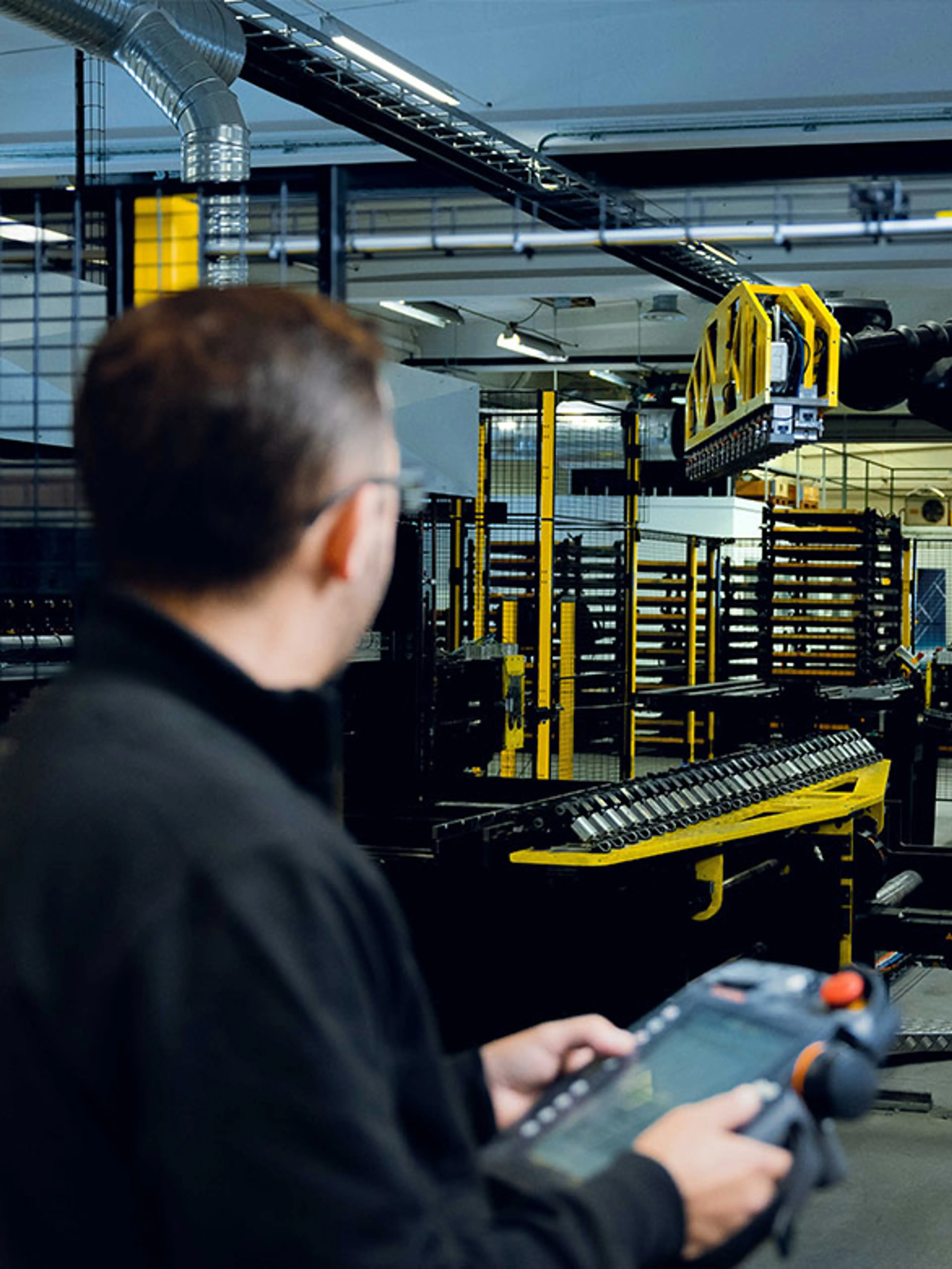 An operator in a black uniform is monitoring an automated industrial system using a handheld control panel. In the background, a robotic lifting mechanism is handling materials, while safety fencing and structured work zones ensure a secure working environment.