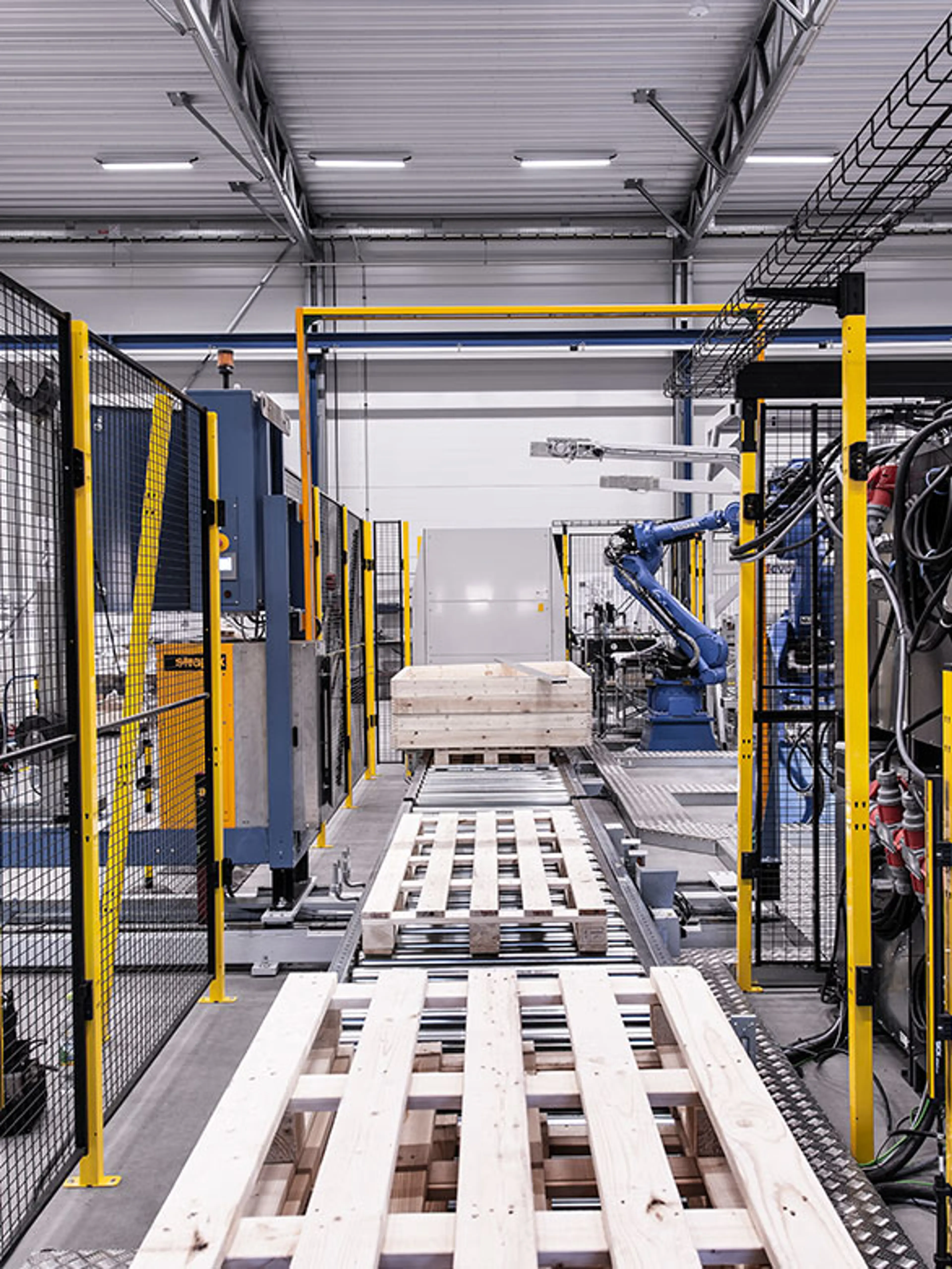 An industrial robotic work cell with protective fencing and automated machinery handling wooden pallets. A blue robotic arm is visible in the background, performing tasks along a conveyor system. Overhead cable trays and safety barriers indicate adherence to robot safety principles.