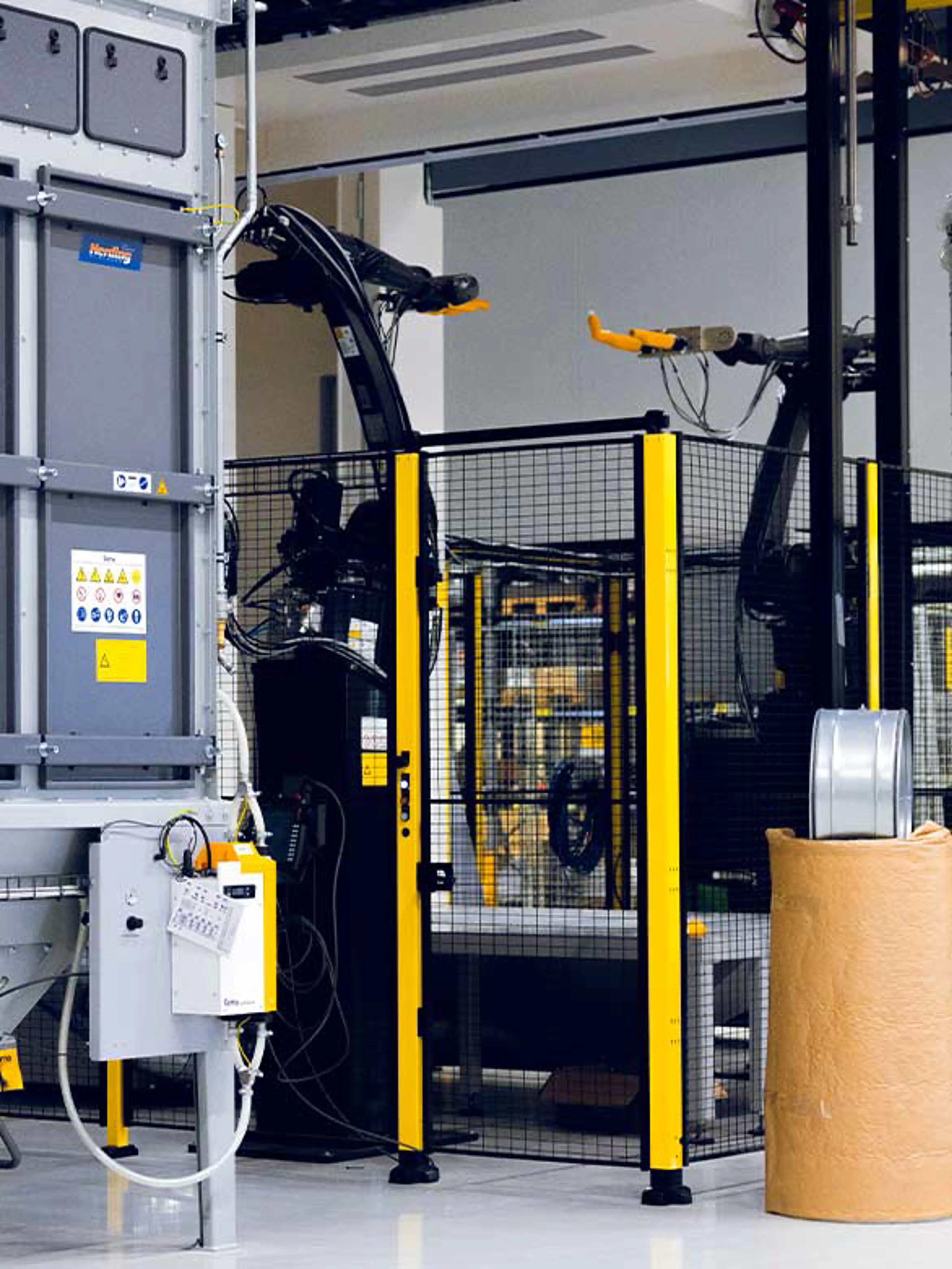 An industrial robotic system enclosed by black and yellow safety fencing. A large machine is visible on the left with control panels and warning labels, ensuring compliance with safety standards. Nearby, a roll of material and a metal component are positioned for processing