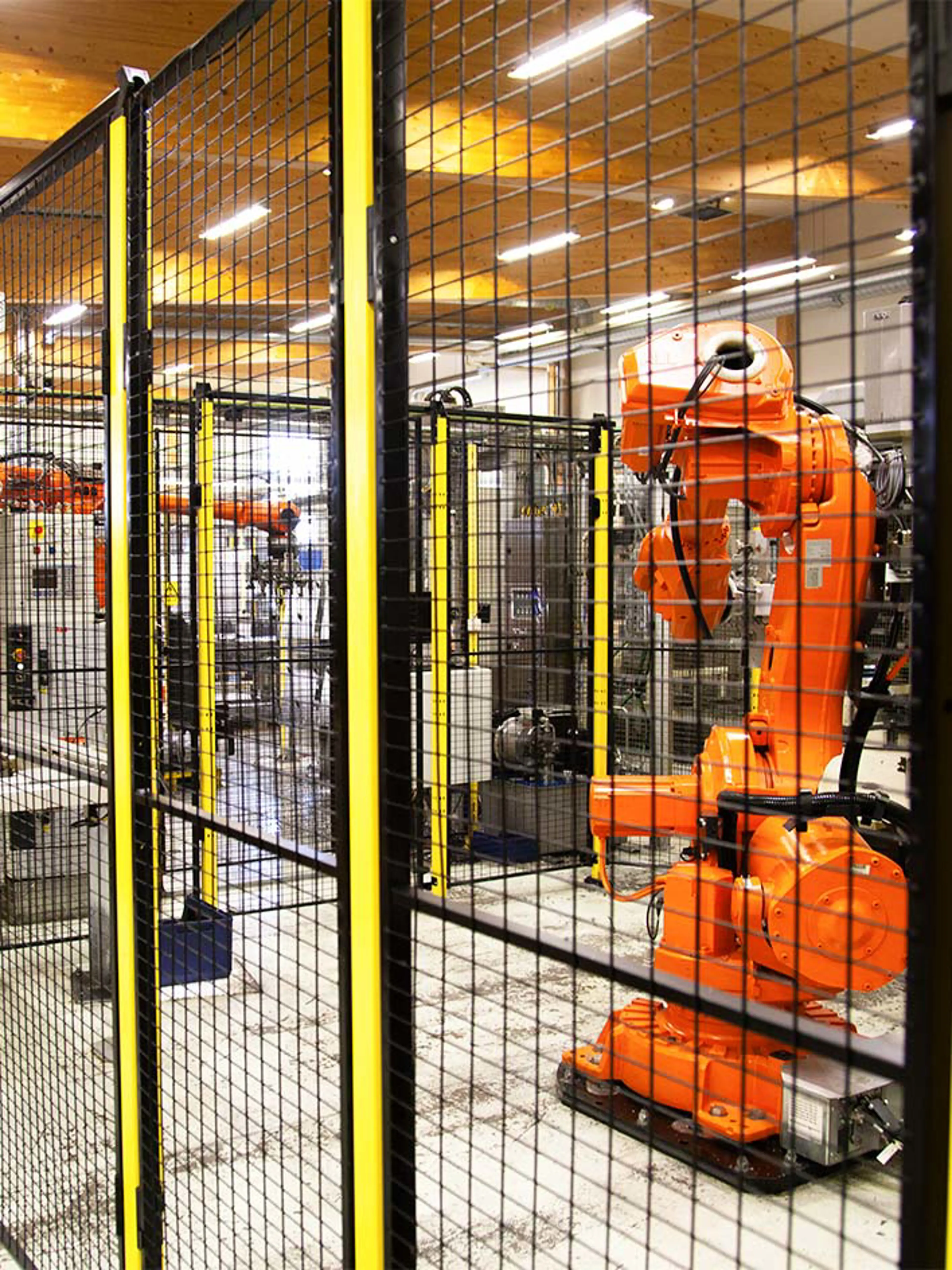 Bright orange industrial robotic arm operating inside a secure, fenced-off automation cell. The black and yellow safety fencing ensures compliance with machine safety regulations, preventing unauthorized access while enabling efficient robotic manufacturing processes. Overhead lighting and a clean workspace highlight a well-organized industrial setting.