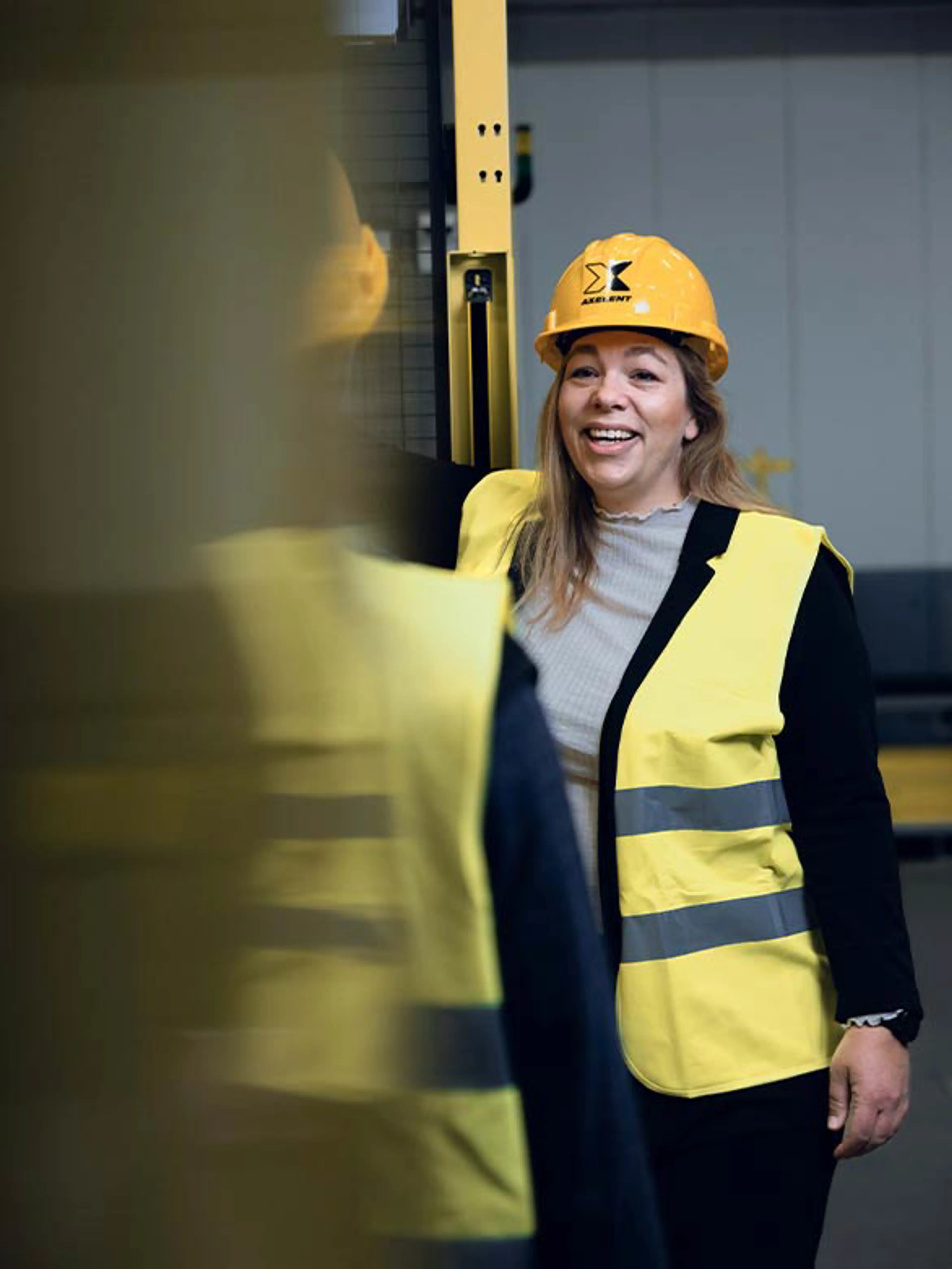 A smiling worker wearing a hard hat and high-visibility vest in an industrial setting, engaging in conversation near safety barriers. The image emphasizes the importance of a positive safety culture and the role of machine safety in creating a secure and efficient work environment.