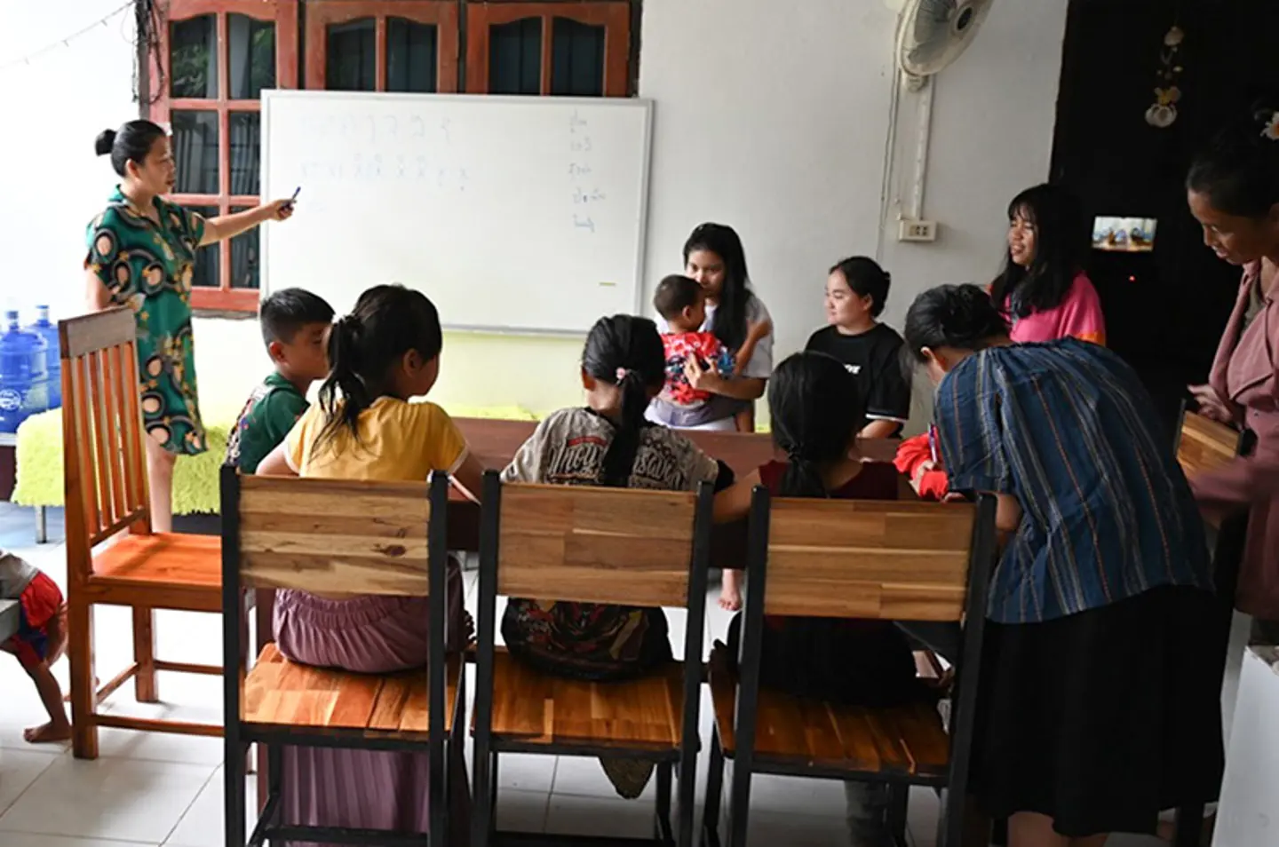 Adult woman teaching at a whiteboard while several girls and caregivers sit together in a classroom at a family home in Laos